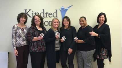 A small group of professionals standing in front of the Kindred at Home logo. 