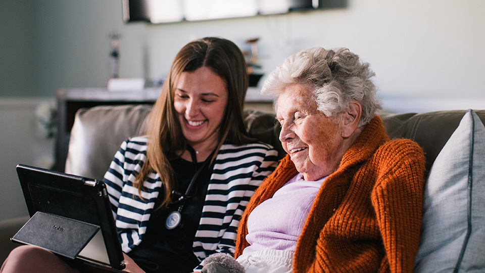 A clinician and a patient seated on a sofa, smiling and looking at a tablet.