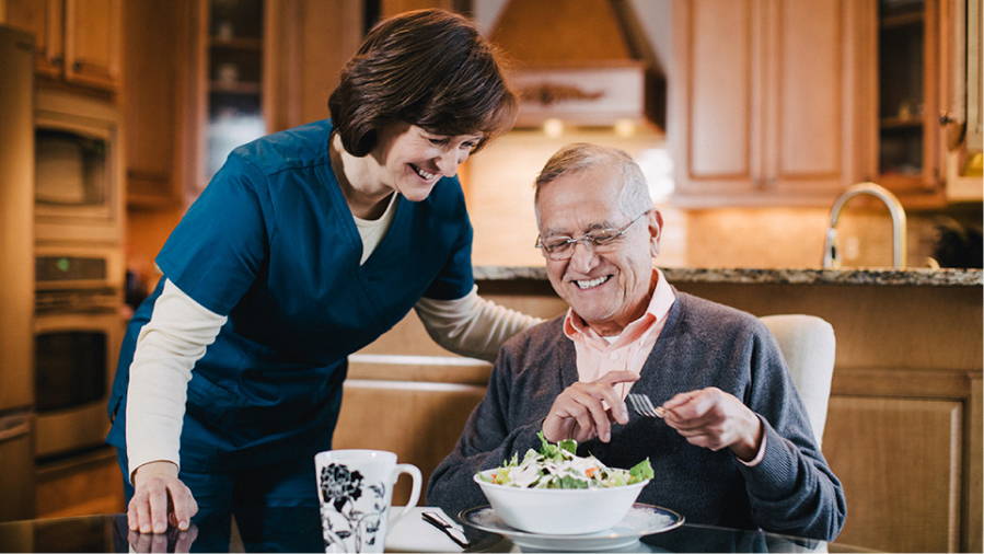 A personal home caregiver smiling beside an elderly patient eating a salad.