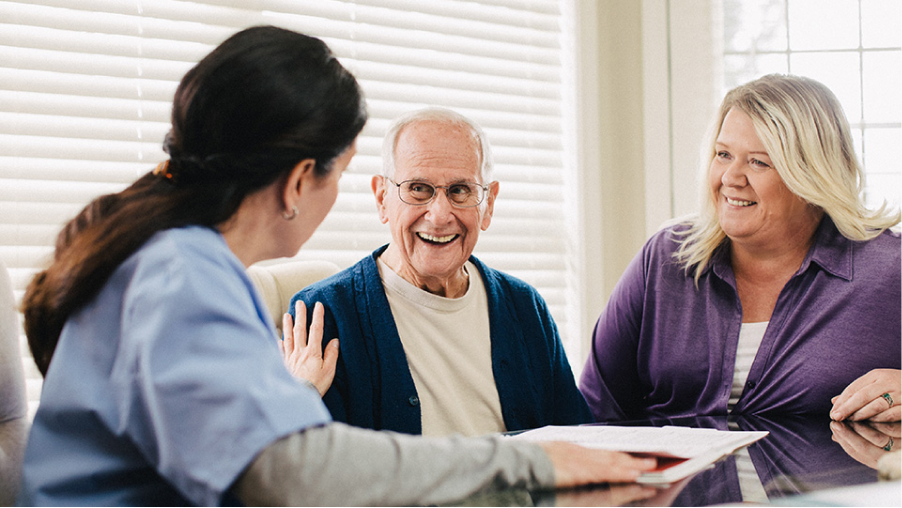 A personal home caregiver gently touching a smiling patient who’s seated beside a family member. 