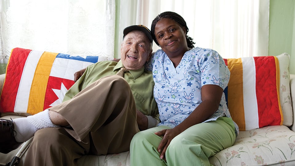 A personal home caregiver and an elderly patient, smiling and seated on a sofa. 