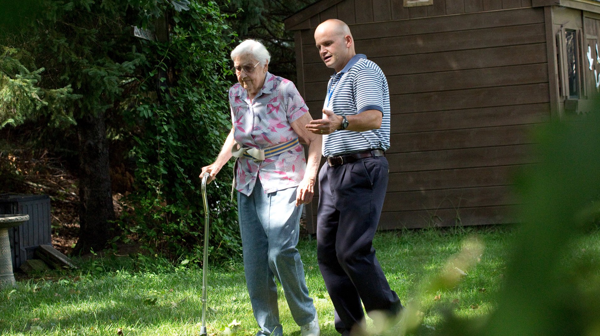 A home health physical therapist helping an elderly patient take an outdoor walk.