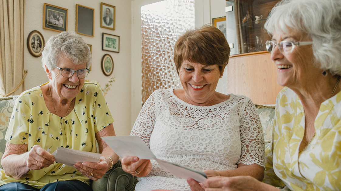 Three elderly women smiling, reading letters and enjoying each other’s company.