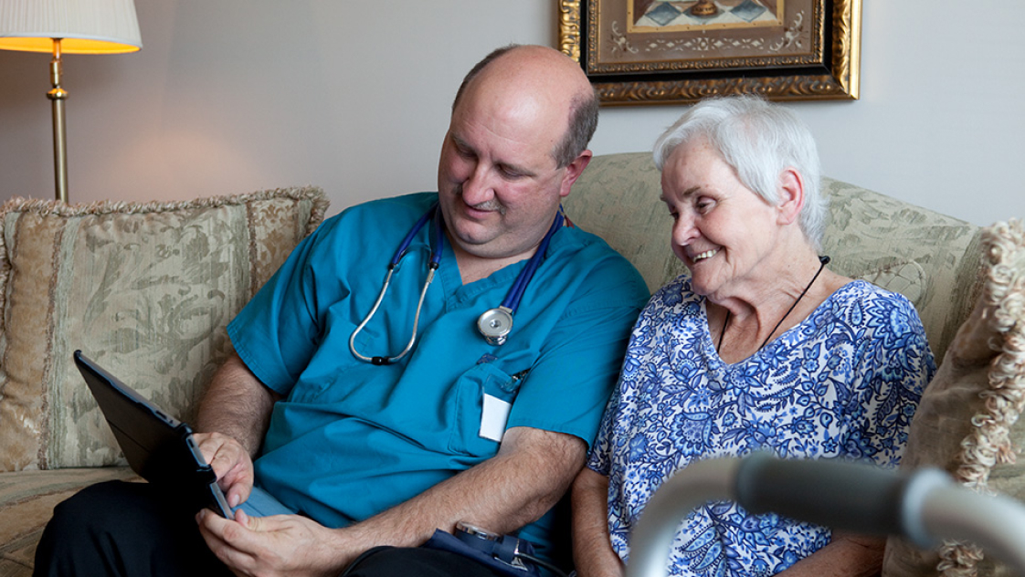 A clinician seated beside an elderly patient on a sofa, explaining medical information on a tablet.