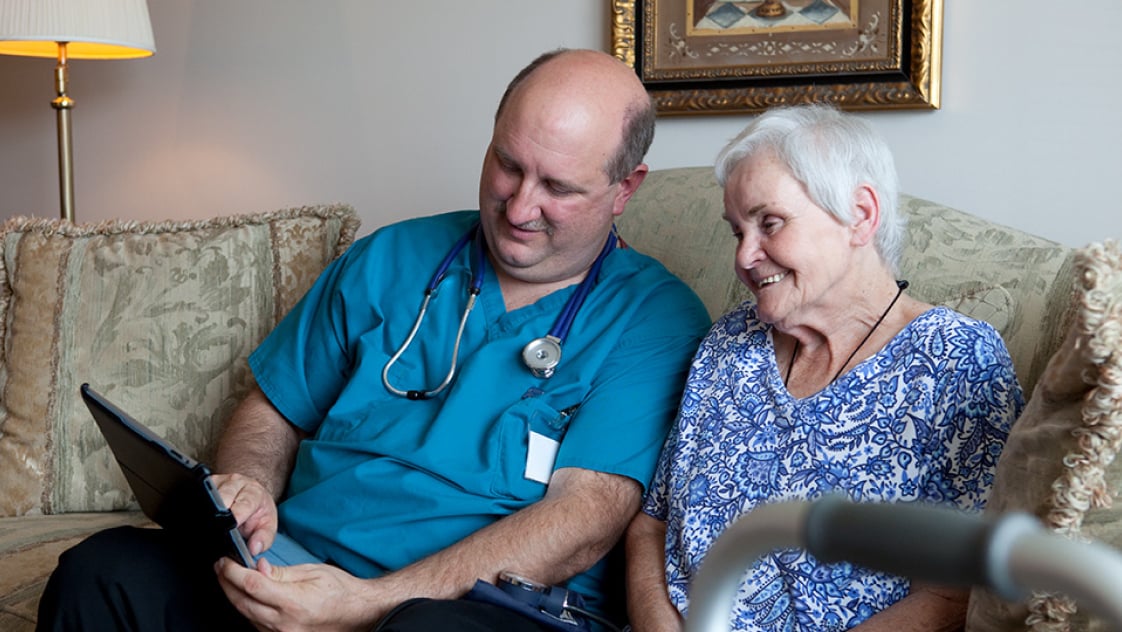 A clinician seated beside an elderly patient on a sofa, explaining medical information on a tablet.