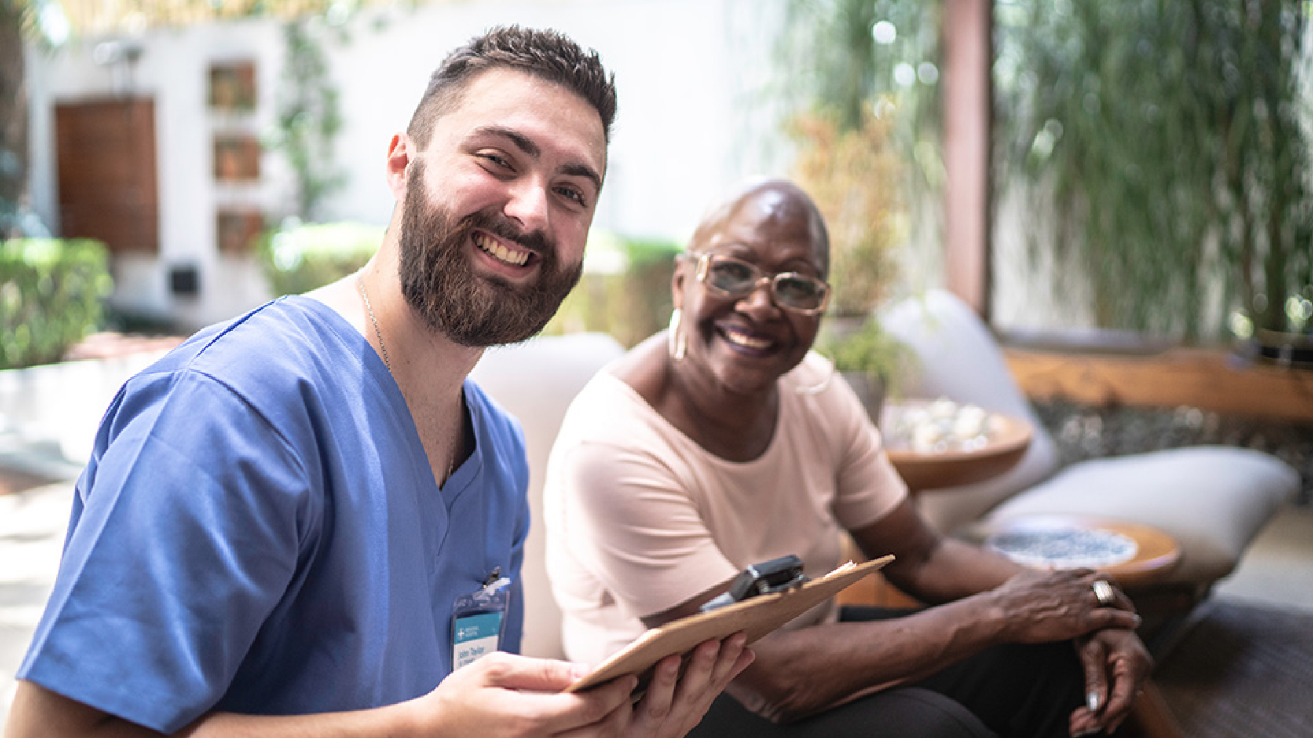 A smiling caregiver holding a clipboard while seated beside a smiling elderly patient.