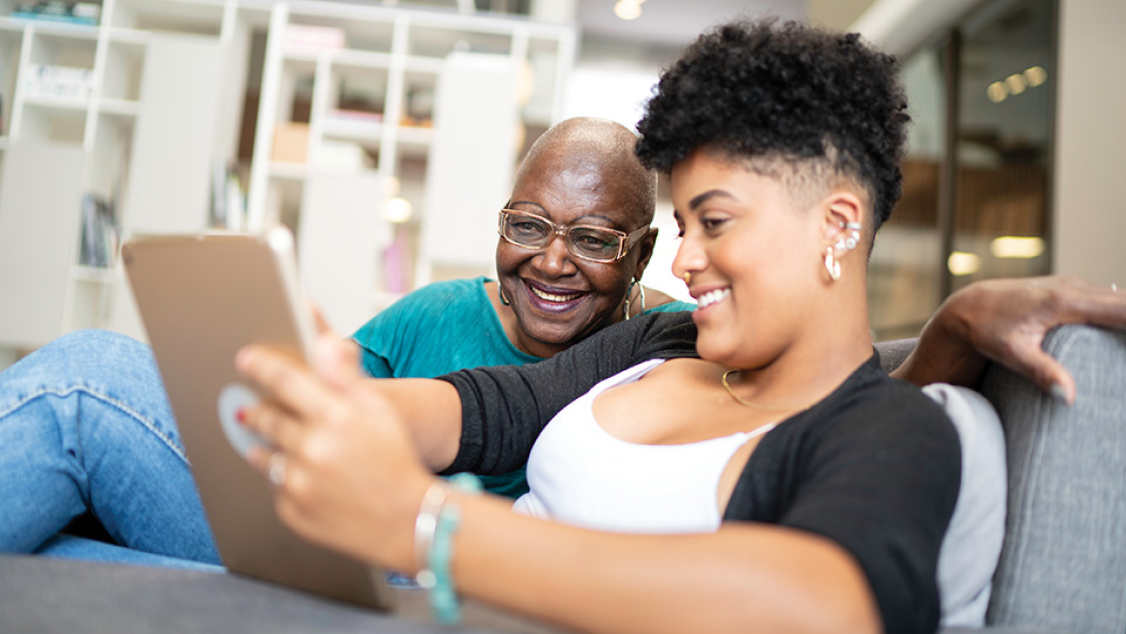 An elderly adult and a family member seated on a sofa, reading information on a tablet.
